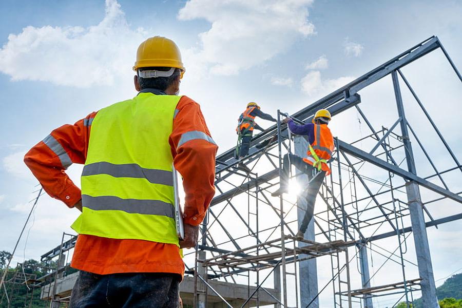 An individual in a yellow safety vest and hard hat watches two other individuals in similar attire climbing scaffolding